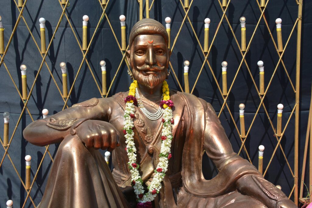 A bronze statue of Shivaji Maharaj adorned with a floral garland, set against a decorative backdrop.